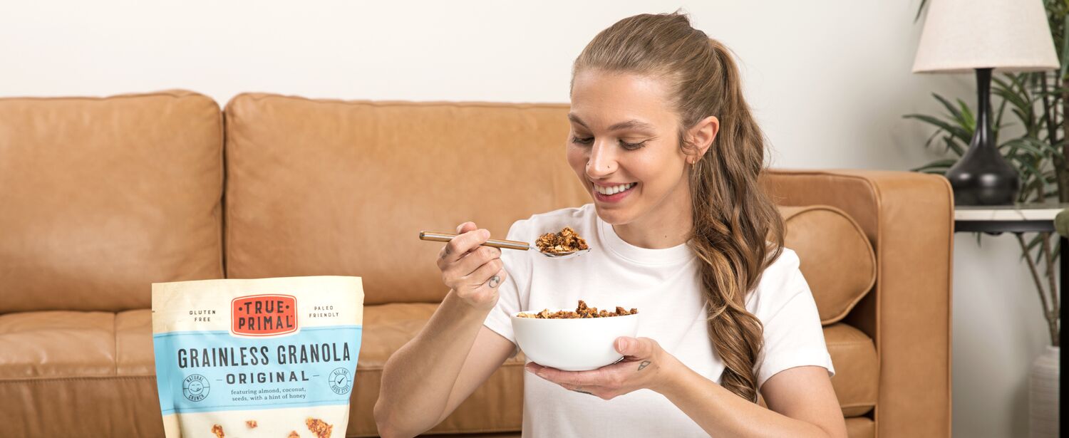 Woman eating True Primal Grainless Granola Original from bowl next to granola pouch